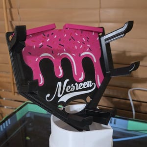 May include: A black and pink phone stand with a donut design. The stand features a pink frosted top with sprinkles and white icing drips. The name "Nesreen" is written in white script on the black background.