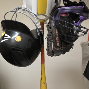 May include: A softball equipment set includes a black helmet with a softball sticker, a black and purple glove, a yellow water bottle with a red softball design, and a softball bat with the word "HOPE". The equipment is hanging on a white rack.