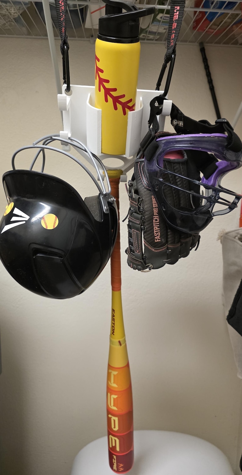May include: A softball equipment set includes a black helmet with a softball sticker, a black and purple glove, a yellow water bottle with a red softball design, and a softball bat with the word "HOPE". The equipment is hanging on a white rack.
