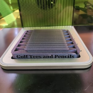 Könnte beinhalten: Ein rechteckiger Behälter mit lila Innenraum und silberner Außenseite. Der Behälter enthält mehrere Golf-Tees. Der Text "Golf Tees and Pencils" ist auf der Seite des Behälters aufgedruckt.
