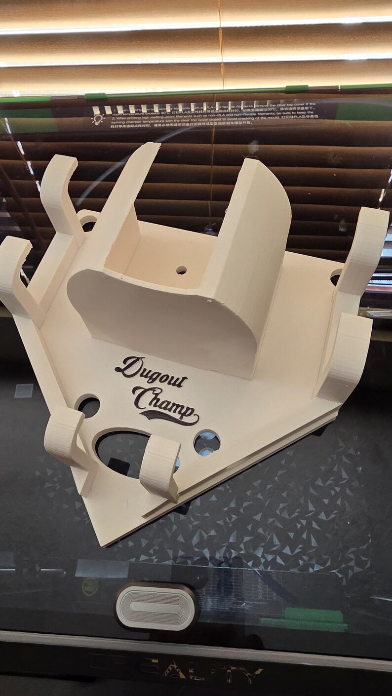 May include: A light beige, 3D-printed baseball dugout display. The display features a home plate shape with the text "Dugout Champ" in a stylized font. It has multiple slots and supports for holding items.