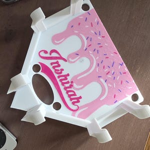 May include: A white phone stand with a pink, dripped frosting design and colorful sprinkles. The stand has the name "Inshirah" in pink script. The stand has several white support arms.