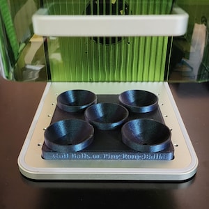May include: A 3D-printed device with five dark blue, bowl-shaped holders. The holders are arranged on a black rectangular base, with the text "Golf Balls or Ping Pong Balls" printed on the base. The device is set inside a larger, green-tinted structure.