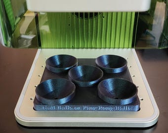 May include: A 3D-printed device with five dark blue, bowl-shaped holders. The holders are arranged on a black rectangular base, with the text "Golf Balls or Ping Pong Balls" printed on the base. The device is set inside a larger, green-tinted structure.