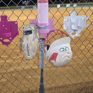 May include: A baseball setup on a chain-link fence. The equipment includes a pink water bottle, a baseball glove, a baseball bat, and a white and red helmet. The bat has a blue and white grip. The background is a baseball field.