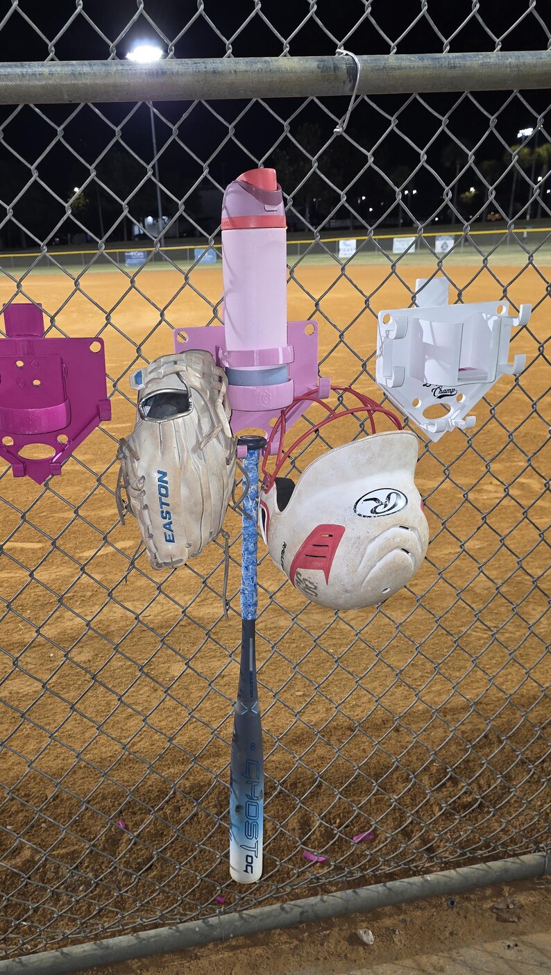 May include: A baseball setup on a chain-link fence. The equipment includes a pink water bottle, a baseball glove, a baseball bat, and a white and red helmet. The bat has a blue and white grip. The background is a baseball field.