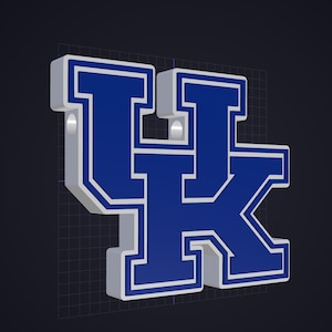 May include: A three-dimensional, block-style letter emblem featuring the letters UK in a bold, dark blue font with a thick white border. The design has a raised, layered appearance with visible side depth and two small circular holes at the top.
