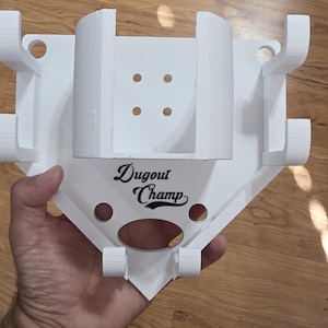May include: A white baseball-shaped holder with the text "Dugout Champ" in black script. The holder has multiple slots and holes, likely for holding baseball bats and other equipment. The design is 3D-printed.