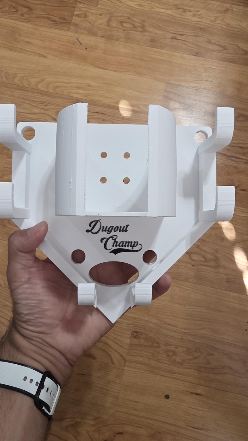 May include: A white baseball-shaped holder with the text "Dugout Champ" in black script. The holder has multiple slots and holes, likely for holding baseball bats and other equipment. The design is 3D-printed.
