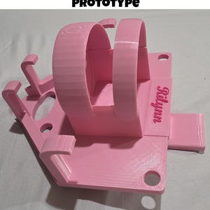 May include: A pink 3D-printed prototype watch stand with the text "Prototype" at the top. The stand features two curved arches for holding the watch and a base with the name "Rilynn" printed on it.