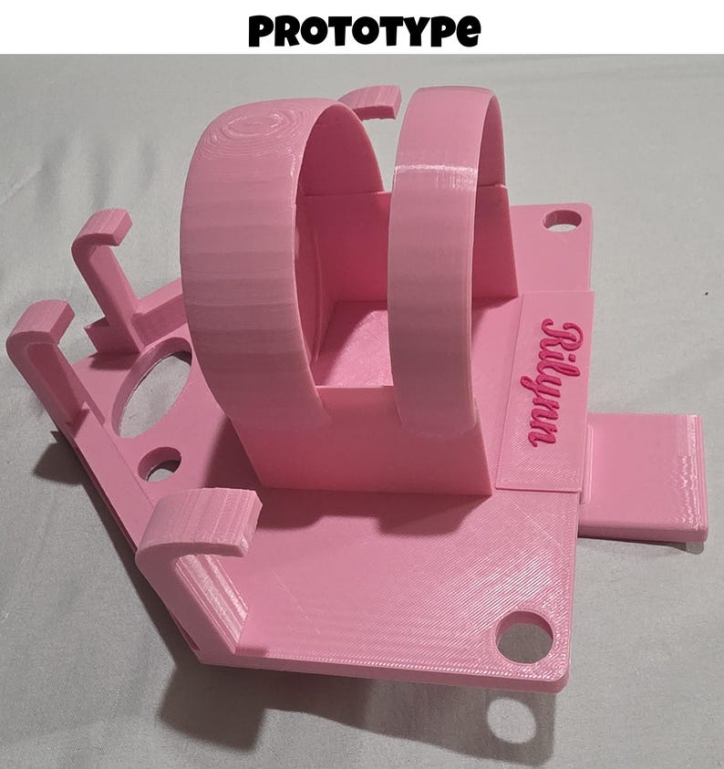 May include: A pink 3D-printed prototype watch stand with the text "Prototype" at the top. The stand features two curved arches for holding the watch and a base with the name "Rilynn" printed on it.