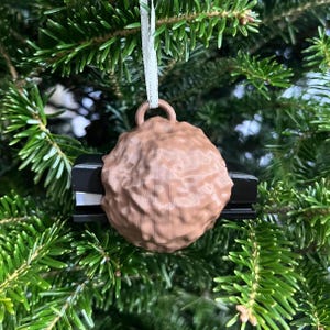 May include: A brown, textured ornament shaped like a stapler, hanging from a white ribbon. The ornament is suspended against a backdrop of green Christmas tree needles. A unique holiday decoration.