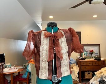 Może przedstawiać: Przezroczysta brązowa bluzka z białym haftem i kokardką. Bluzka ma bufiasty rękaw i jest prezentowana na manekinie.