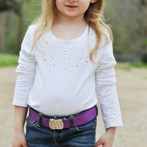 Op de afbeelding: Een jong persoon draagt een witte top met lange mouwen versierd met gouden en zilveren studs, gecombineerd met een blauwe jeans en een paarse glitterriem. De riem heeft een zilveren gesp met de letter "A".