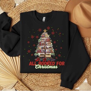 Peut inclure: Sweat-shirt noir avec un motif d'arbre de Noël fait de livres, surmonté d'une étoile. L'expression "All Booked for Christmas" est imprimée en rouge et vert. Des flocons de neige rouges et verts sont dispersés autour du motif de l'arbre.