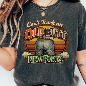 Puede incluir: Camiseta gris oscuro con el texto "Can't Teach an OLD BUTT New Farts" en un diseño retro. El gráfico presenta una vista trasera de una persona con vaqueros azules. La camiseta es una prenda informal.