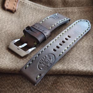 May include: A pair of brown leather watch straps with green stitching. One strap has a silver buckle, and the other has several adjustment holes. The text "TYMA 55-75 WAR IS HELL" is embossed on one strap.