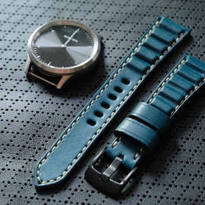 May include: A silver Garmin watch with a black face and a teal leather watch band with white stitching. The watch band has a black buckle. The watch and band are on a dark gray textured surface.