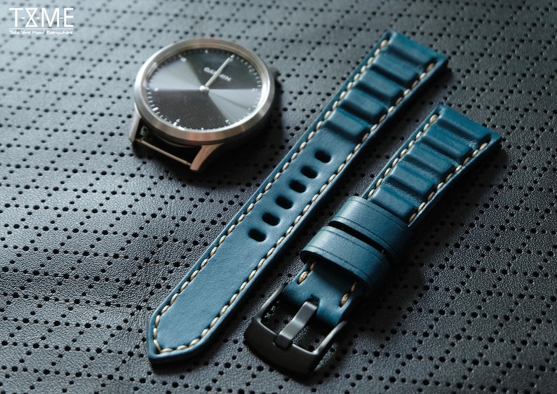 May include: A silver Garmin watch with a black face and a teal leather watch band with white stitching. The watch band has a black buckle. The watch and band are on a dark gray textured surface.