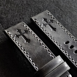 May include: A pair of dark gray leather watch straps featuring a debossed cross emblem on each piece. The straps are constructed with light-colored stitching along the edges and include a sliding leather keeper loop and pre-punched buckle holes.