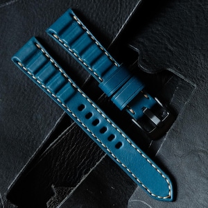 May include: Teal blue watch strap with white stitching. The leather strap features a textured design and a silver-tone buckle. The strap is shown on a dark leather surface. The watch strap is a replacement band.