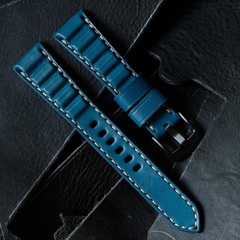 May include: Teal blue watch strap with white stitching. The leather strap features a textured design and a silver-tone buckle. The strap is shown on a dark leather surface. The watch strap is a replacement band.