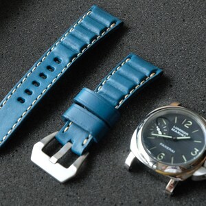 May include: A blue leather watch strap with white stitching and a silver buckle is displayed next to a silver watch with a black face. The watch face has white numerals and hands. Two silver pins are also visible.