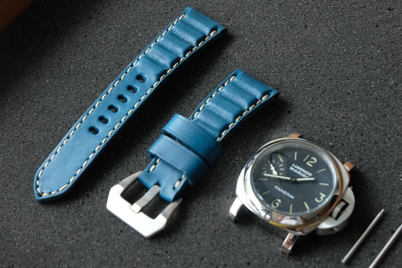 May include: A blue leather watch strap with white stitching and a silver buckle is displayed next to a silver watch with a black face. The watch face has white numerals and hands. Two silver pins are also visible.