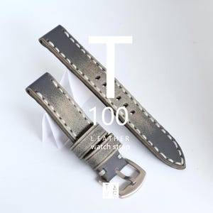 May include: Two-piece gray leather watch strap with white stitching. The strap has a silver buckle and the text "T 100 LEATHER watch strap". The watch strap is displayed on a white background.