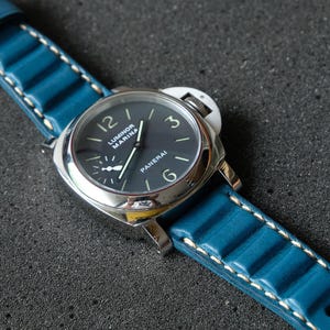 May include: A close-up of a wristwatch with a dark blue face and a silver case. The watch has a blue leather band with white stitching. The watch face displays the brand name "Panerai".