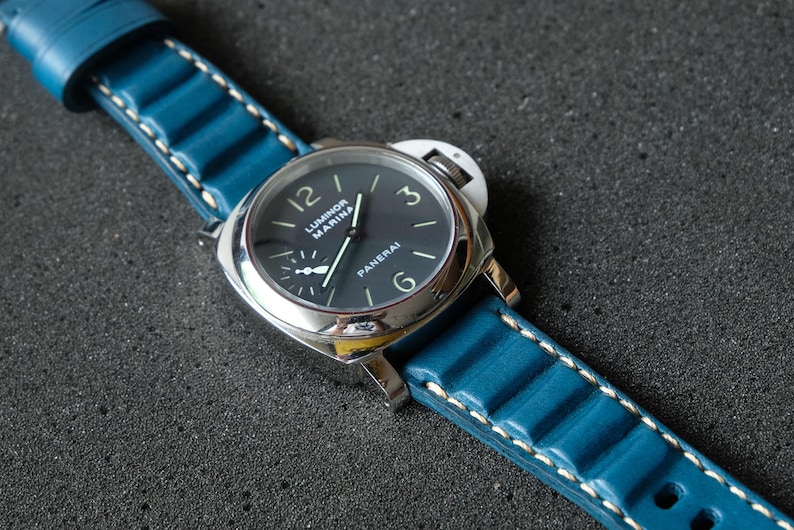 May include: A close-up of a wristwatch with a dark blue face and a silver case. The watch has a blue leather band with white stitching. The watch face displays the brand name "Panerai".
