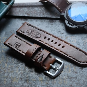 May include: A brown leather watch strap with a silver buckle and white stitching. The strap has the text "Y.M.E. 85-75 WAR IS HELL" and a logo. A black watch with a blue face is also visible.