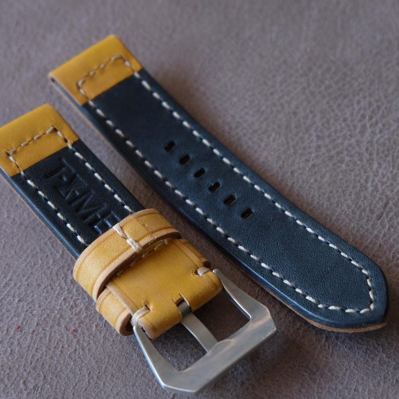 May include: A close-up of two watch straps. The straps are navy blue with yellow accents and white stitching. One strap has a silver buckle. The other strap has the letters "J-XME" embossed on it.