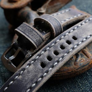 Può includere: Primo piano di un cinturino per orologio in pelle grigia invecchiata con cuciture bianche. Il cinturino presenta una fibbia argentata e diversi fori di regolazione. La pelle ha un aspetto usurato e vintage, che suggerisce un design artigianale. Il cinturino è un ricambio per un orologio.