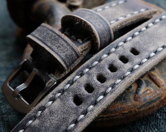 May include: Close-up of a distressed gray leather watch strap with white stitching. The strap features a silver buckle and multiple adjustment holes. The leather has a worn, vintage appearance, suggesting a handcrafted design. The watch band is a replacement part for a watch.