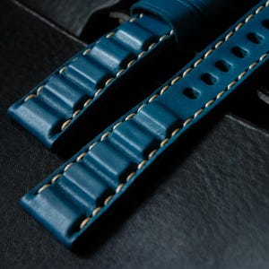 May include: Close-up of two teal blue leather watch straps. The straps feature a textured design with raised ridges and are stitched with light beige thread. One strap has oval holes. The background is a dark, textured surface.
