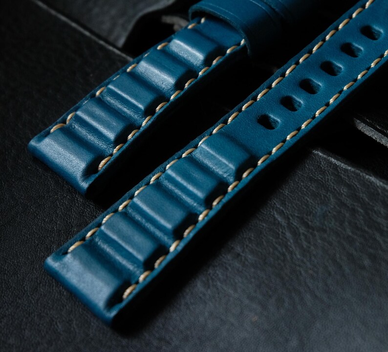 May include: Close-up of two teal blue leather watch straps. The straps feature a textured design with raised ridges and are stitched with light beige thread. One strap has oval holes. The background is a dark, textured surface.
