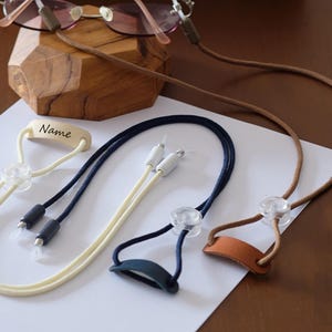 May include: Eyeglass chains in brown, navy, and cream colors. One chain has a small wooden piece with the word "Name" engraved. The chains have clear, adjustable loops to attach to glasses. The chains are designed to keep glasses secure.