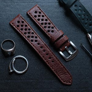 May include: Brown leather watch straps with perforated holes and a silver buckle. The straps are textured and have a classic design. A silver ring is also visible.