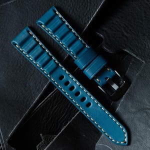 May include: Two-piece teal watch strap with white stitching. The strap features a textured design and a silver-tone buckle. The watch band is displayed on a dark, textured surface.