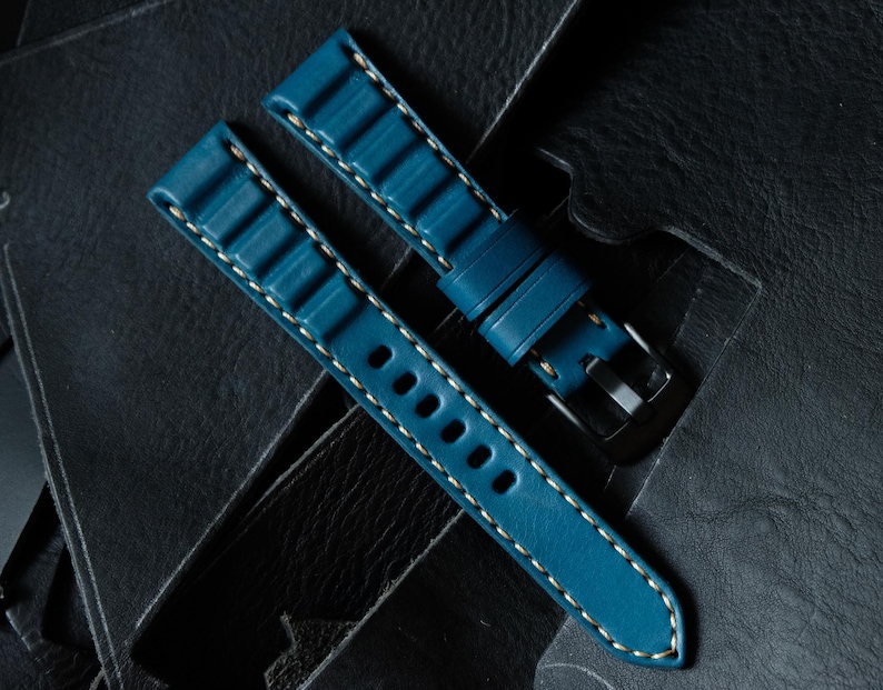 May include: Two-piece teal watch strap with white stitching. The strap features a textured design and a silver-tone buckle. The watch band is displayed on a dark, textured surface.