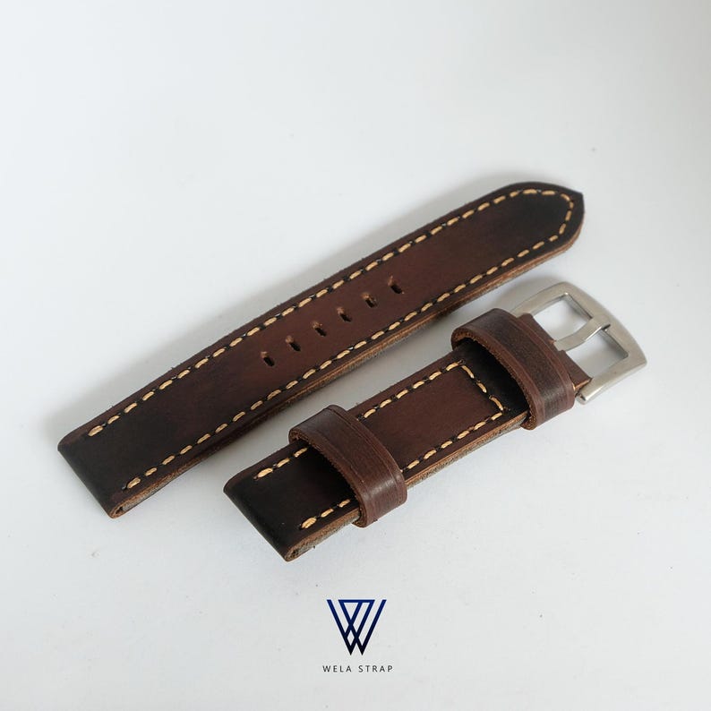 May include: A brown leather watch strap with white stitching and a silver buckle. The strap has a distressed appearance and the text "WELA STRAP" is visible.
