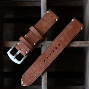 May include: A brown leather watch strap with a silver buckle. The strap has white stitching and several holes for adjustment. The leather has a distressed look, and the strap is displayed on a wooden surface.