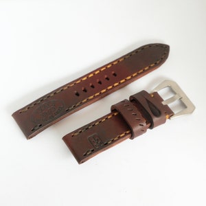 May include: A dark brown leather watch strap featuring thick tan stitching and a silver-toned metal buckle. The strap is embossed with the text T.Y.M.E 55-75 WAR IS HELL and a small symbol. The leather has a smooth finish with visible edge detailing.