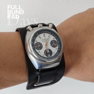Puede incluir: Un reloj cronógrafo vintage con caja plateada y esfera en blanco y negro, con agujas naranjas y visualización de la fecha. El reloj está montado en una amplia pulsera de cuero negro. El texto "FULL BUND PAD Days" es visible.