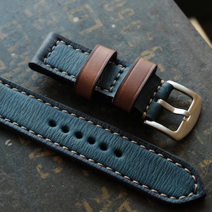 May include: Two-piece watch strap in blue leather with brown leather accents. The strap features cream-colored stitching and a silver-colored buckle. The watch band is designed for a classic and stylish look.