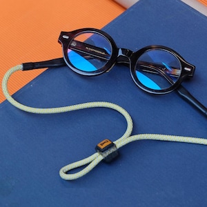 Pode incluir: Óculos redondos, com armação tartaruga e lentes azuis, sobre uma superfície azul. Um cordão verde claro e amarelo está preso aos óculos, com um pequeno conector quadrado castanho e preto. Os óculos têm um estilo clássico e vintage.