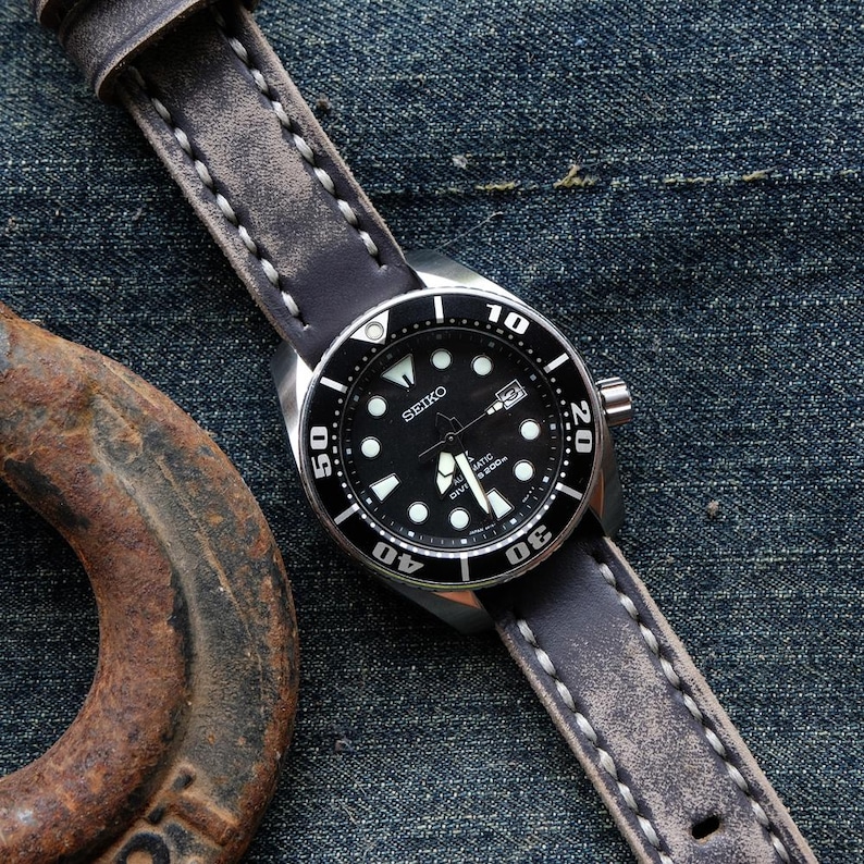 May include: A black Seiko dive watch with a black bezel and a black dial, featuring white luminous markers and hands. The watch has a gray leather strap with white stitching. The watch is displayed on a denim background.