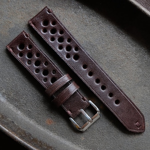 May include: A dark brown leather watch strap featuring a perforated rally-style design with multiple circular cutouts. The strap includes two matching leather keepers and a polished silver-tone metal pin buckle for secure fastening.
