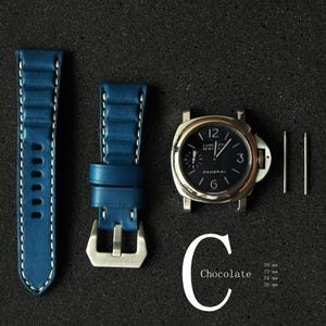 May include: A watch face with a black dial and silver casing, accompanied by a blue leather watch strap with white stitching and a silver buckle. Two silver pins are also included. The word "Chocolate" is written in white.
